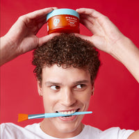 Model with curly hair holding Halo Hydrator Scalp Mask and Applicator on red background.